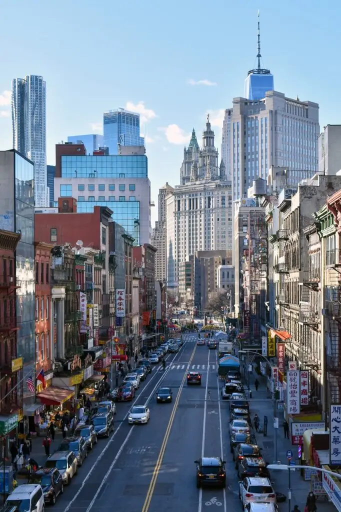 Busy Chinatown street in Lower Manhattan lined with storefronts and parked cars, with high-rise towers in the distance