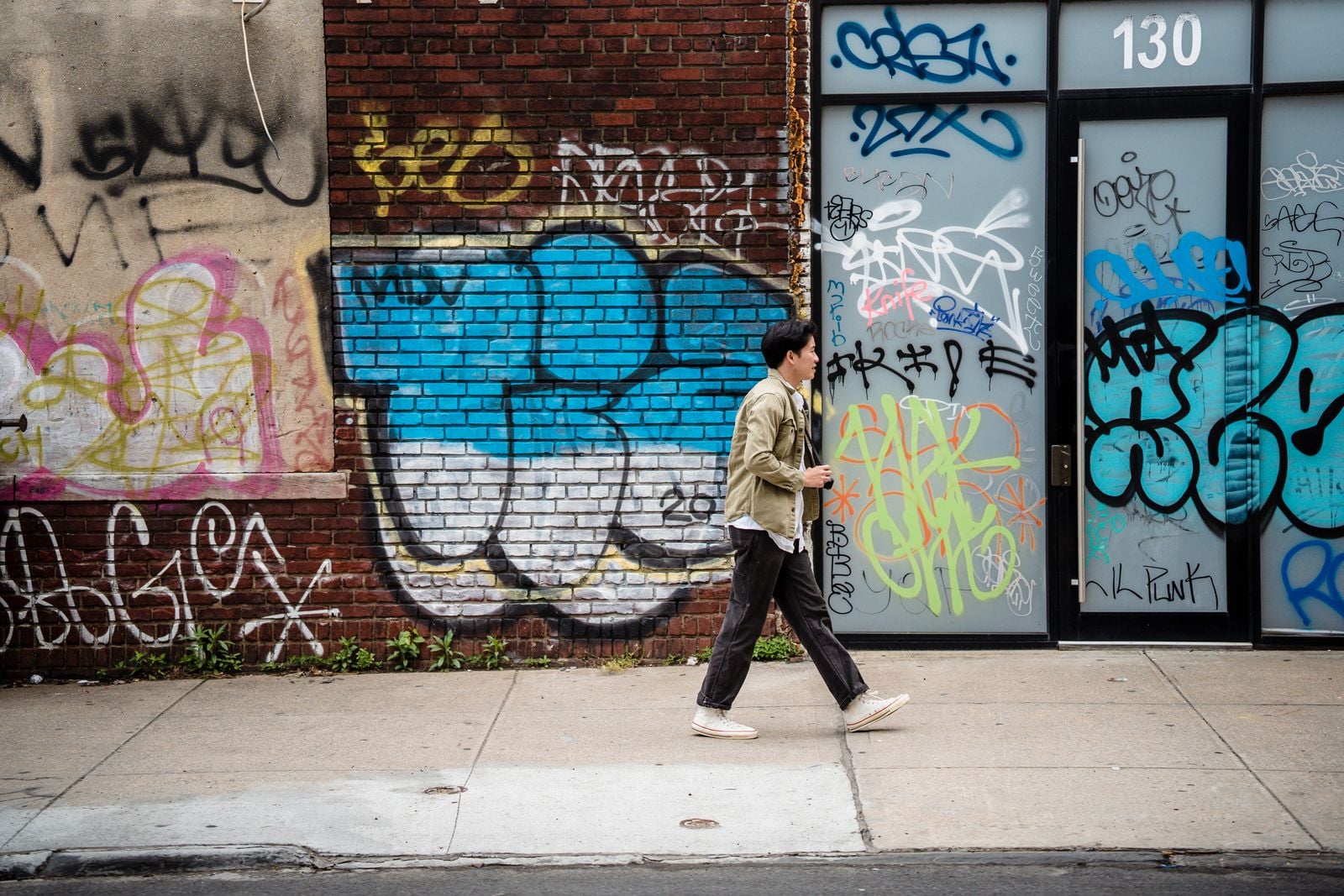 Man in a casual jacket and everyday pants walking past a graffiti wall on a city sidewalk, the kind of streetwear look concealed carry clothing should blend into