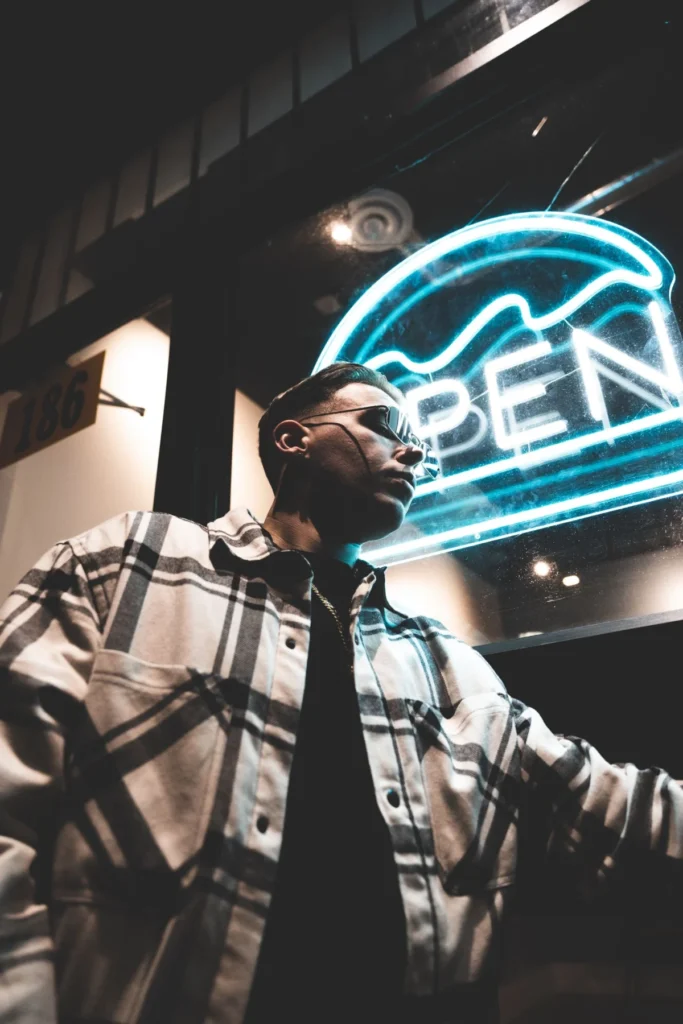 A man wearing an open black-and-white check overshirt over a dark T-shirt, standing under a neon sign at night